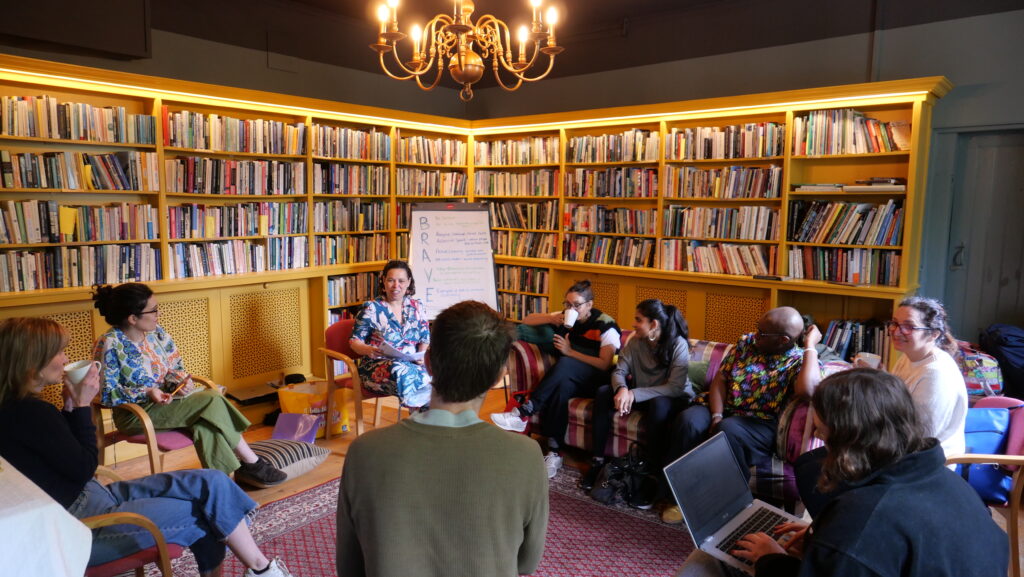 The image is set in a small library room, with several people on chairs discussing the organisation.