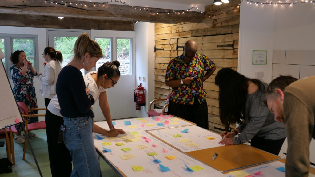Several SPD board members and employees are in a barn, standing over a table full of different coloured notes, they are adding to the notes, and chatting.